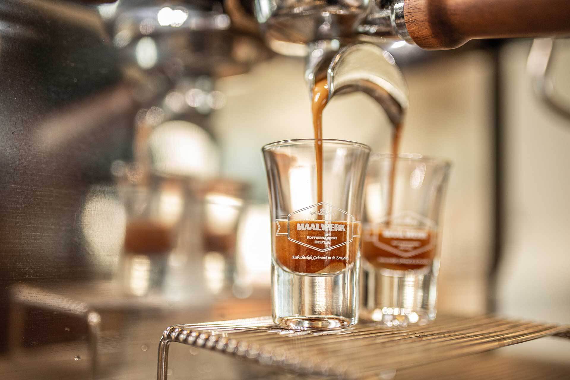 Twee espressoglazen met maalwerk logo onder een barista koffiemachine